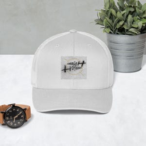 Könnte beinhalten: Weiße Trucker-Cap mit gebogenem Schirm und Mesh-Rückseite. Auf der Vorderseite befindet sich ein quadratischer Aufnäher mit dem Text "Werrich Horizon" in schwarzer Schrift und einem goldenen Kreisdesign. Ein modisches Accessoire.