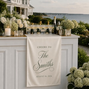 May include: A cream-colored fabric table runner draped over a white bar, featuring the black printed text CHEERS TO The Smiths AUGUST 16, 2026. The runner is surrounded by white floral arrangements, clear glass stemware, and bottles of champagne.
