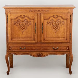 Puede incluir: Un mueble de madera marrón claro con dos puertas y un cajón. Las puertas presentan tallas ornamentadas y el cajón tiene un tirador decorativo. El mueble tiene patas curvas y un adorno detallado. Es un mueble.