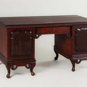 May include: A dark brown wooden desk with a rectangular top and ornate detailing. The desk features a single drawer and decorative panels on the sides. The desk has curved legs and a classic design.