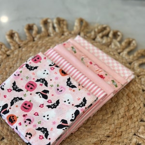 May include: A stack of five folded fabric squares featuring various pink and white patterns. The top piece displays a Halloween-themed print with pink pumpkins, white ghosts, and black bats on a light pink background, followed by striped and checkered fabrics.