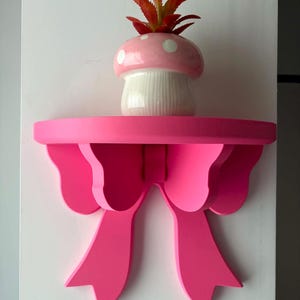 Puede incluir: Una estantería de pared de color rosa brillante con forma de lazo y superficie superior plana. Sobre la estantería hay una pequeña maceta de cerámica con forma de seta, con sombrero rosa de lunares blancos y base blanca estriada, con una planta naranja.
