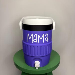 以下が含まれることがあります： 白い文字で「MAMA」と書かれた紫色のウォータークーラー。クーラーには白い蓋とハンドル、黒いバンド、白い蛇口が付いています。小さな緑色のスツールの上にあります。