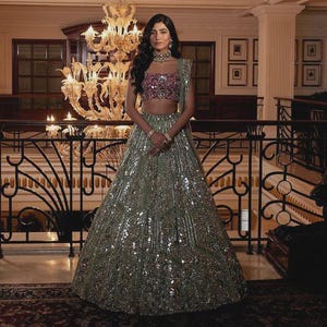 May include: A woman in a formal gown stands in a grand hall. The gown is a light green color and covered in sequins. The top is a pink and purple sequined crop top. A large chandelier hangs above.