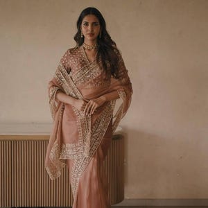 May include: A dusty rose saree featuring a sheer fabric with intricate gold embroidery along the borders and bodice. The garment includes a matching blouse with a V-neckline and detailed embellishments, paired with a gold choker necklace.