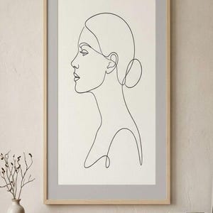 Könnte beinhalten: Gerahmter Kunstdruck mit einer minimalistischen Strichzeichnung des Profils einer Frau. Das Kunstwerk verwendet schwarze Linien auf hellem Hintergrund, gerahmt in einem hellen Holzrahmen. Das Haar der Frau ist zu einem Dutt gebunden.