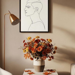 Könnte beinhalten: Eine gerahmte Schwarz-Weiß-Strichzeichnung eines Männerprofils hängt über einem Tisch. Links ist eine goldene Lampe. Eine weiße und braune Vase enthält einen Strauß aus orangefarbenen, gelben und roten Blumen. Herbstblätter liegen auf dem Tisch verstreut.