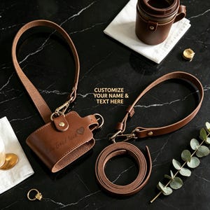 Può includere: Una collezione di accessori in pelle marrone, tra cui una custodia per telefono con cinturino, un portabicchieri e un cinturino arrotolato. La custodia del telefono è incisa con "Lily Good luck" e un cuore. È visibile anche il testo "Customize your name & text here".