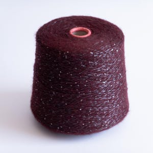 Op de afbeelding: Een conische klos diepbordeauxrood garen met een pluizige, geborstelde textuur. Het garen is verweven met dunne, reflecterende zilveren metallic draden die een subtiele, verspreide schittering creëren over het oppervlak van de donkerrode vezel.