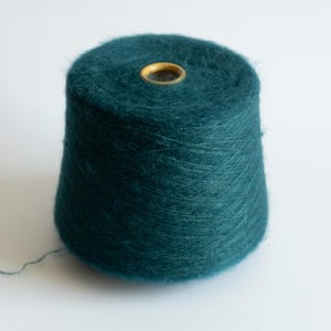 May include: A teal-colored yarn cone with a cylindrical shape and a central hole. The yarn has a soft, fuzzy texture, and a strand of yarn extends from the cone. The cone sits on a white surface.