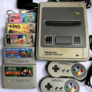 May include: A Nintendo Super Famicom console with several game cartridges and two controllers. The console is gray with a dark gray top and features power, eject, and reset buttons. The game cartridges include Donkey Kong 2 and Donkey Kong.