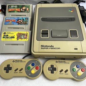 Könnte beinhalten: Eine beige Nintendo Super Famicom Konsole mit drei Spielkassetten und zwei Controllern. Die Kassetten sind mit "Donkey Kong", "Donkey Kong 2" und "Super Mario World" beschriftet. Die Controller haben farbige Knöpfe und ein Steuerkreuz.