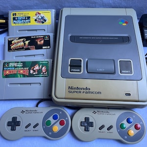Puede incluir: Una consola de videojuegos Nintendo Super Famicom de color beige y gris con dos mandos a juego, un adaptador de corriente y tres cartuchos de juego titulados Super Mario Kart, Street Fighter II y Super Donkey Kong, con etiquetas coloridas.
