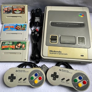 May include: A Nintendo Super Famicom game console with two controllers, a power adapter, and three Donkey Kong game cartridges. The console is light grey with a dark grey centre, featuring power, eject, and reset buttons and a power indicator light.