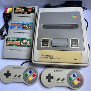 Puede incluir: Una consola Nintendo Super Famicom con dos mandos y tres cartuchos de juego. La consola es gris y beige, con las palabras "Nintendo SUPER FAMICOM" impresas en la parte delantera. Los mandos son grises con botones de colores. Los cartuchos de juego incluyen "Street Fighter II" y dos juegos de "Donkey Kong".