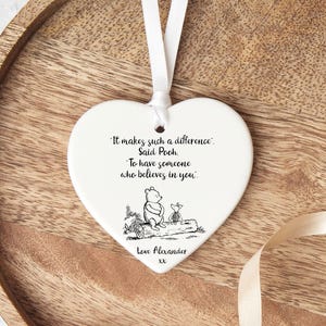 May include: A white heart-shaped ornament with a ribbon. The ornament features a Winnie the Pooh illustration and the quote, "It makes such a difference. Said Pooh. To have someone who believes in you." The text "Love Alexander" is also included.