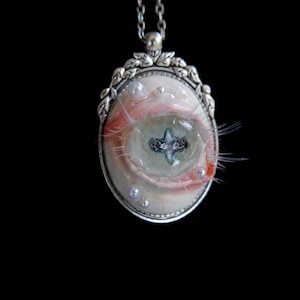 Op de afbeelding: Een ovale hanger aan een ketting met een realistisch vormgegeven oog met roze oogleden, lange wimpers en een kruisvormige pupil. Het oog is gevat in een gedecoreerde, zilverkleurige metalen omlijsting die is versierd met kleine witte parels.