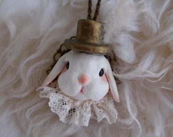 Puede incluir: Un collar con colgante caprichoso que presenta una cabeza de conejo blanco con un pequeño sombrero de copa dorado y un cuello de encaje. El conejo tiene detalles rosas en la nariz y las orejas, y ojos negros. La cadena es de color bronce.