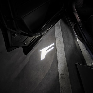 May include: A white illuminated "F" logo projected onto a concrete surface, likely from a car's door. The image is dark, with the car door and interior visible. The logo is bright against the dark background.