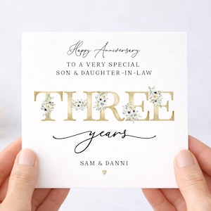 May include: A square white anniversary card featuring the text Happy Anniversary TO A VERY SPECIAL SON & DAUGHTER-IN-LAW. The word THREE is printed in large gold letters decorated with floral illustrations. Below is the text years and SAM & DANNI.
