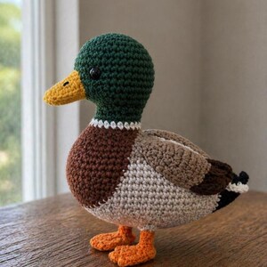 May include: A crocheted mallard duck toy featuring a dark green head, a yellow beak, a brown chest, and a light beige body. The wings are light brown with dark brown and black accents at the tail, and the feet are bright orange. The duck has small black eyes.