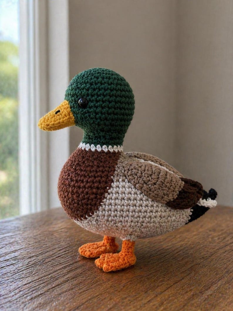 May include: A crocheted mallard duck toy featuring a dark green head, a yellow beak, a brown chest, and a light beige body. The wings are light brown with dark brown and black accents at the tail, and the feet are bright orange. The duck has small black eyes.