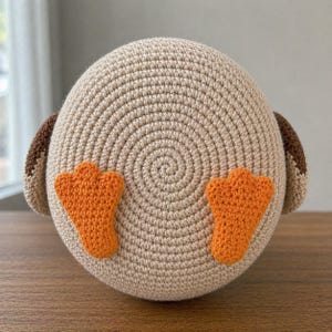 May include: A round, beige crocheted plush toy featuring a spiral stitch pattern on the front. It has two bright orange, webbed, three-toed feet attached to the lower center and two dark brown, rounded, wing-like appendages on the sides.
