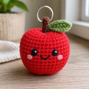May include: A red crocheted apple keychain featuring a brown stem, a green leaf, and a smiling face with black eyes and pink cheeks. The apple is round and textured with yarn stitches, topped with a silver metal ring for attachment purposes.