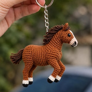 May include: A brown crocheted horse keychain with a silver keyring and chain. The horse has a dark brown mane and tail, white markings on its face, and black and white hooves. The keychain is held up against a blurred green background.