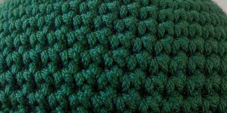 May include: A dark green crocheted fabric featuring a dense, uniform stitch pattern. The texture is created by interlocking loops of yarn, forming a consistent, repeating grid of V-shaped stitches across the entire surface of the material.