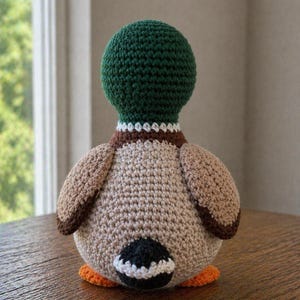 May include: A crochet mallard duck toy featuring a dark green head, a white neck ring, a brown chest, and a tan body. The duck has brown wings, a black tail patch with a white border, and bright orange feet, all crafted with visible yarn stitches.