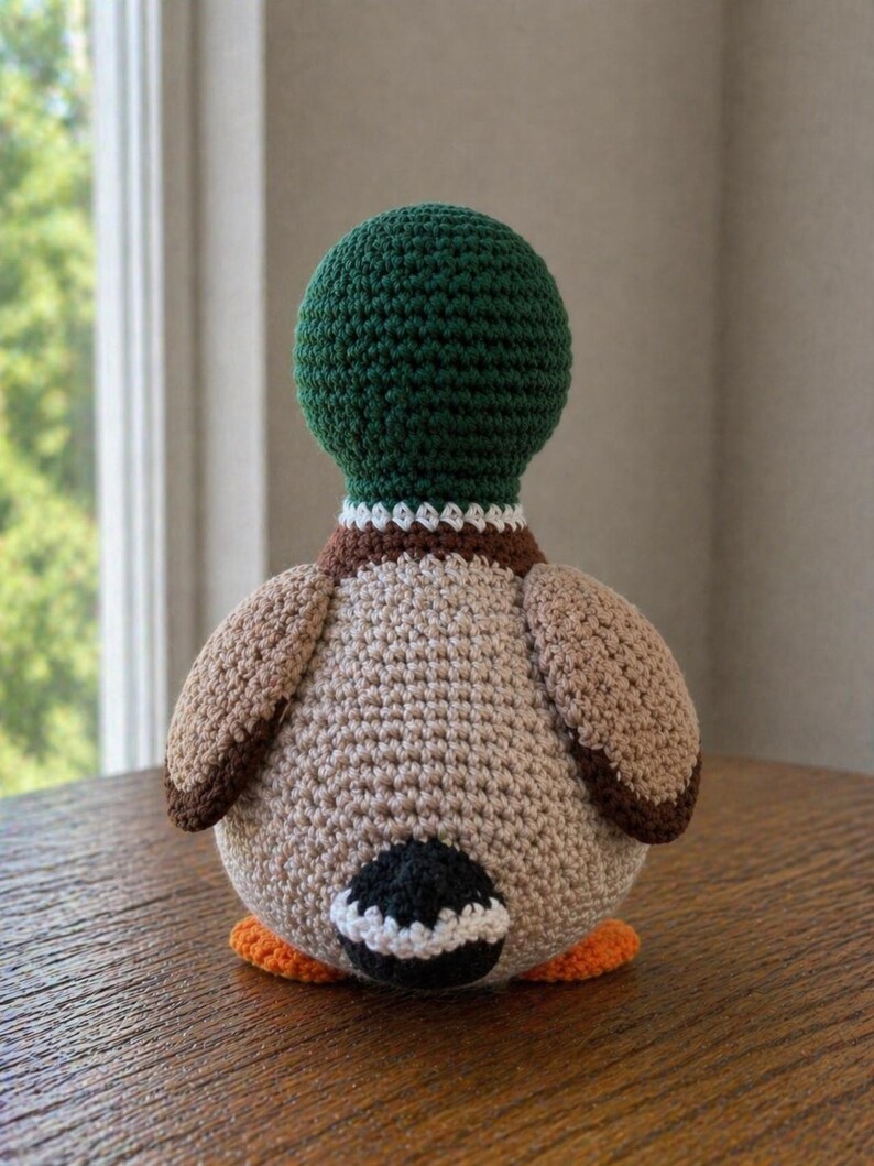 May include: A crochet mallard duck toy featuring a dark green head, a white neck ring, a brown chest, and a tan body. The duck has brown wings, a black tail patch with a white border, and bright orange feet, all crafted with visible yarn stitches.