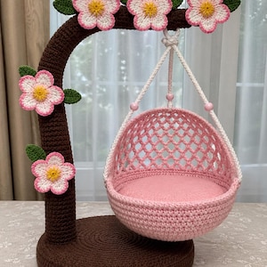 Puede incluir: Un columpio para gatos hecho a mano con ganchillo en tonos rosa y marrón. El columpio tiene una base redonda y un soporte curvo adornado con flores de ganchillo. La cesta colgante tiene un diseño de enrejado y un cojín rosa suave.