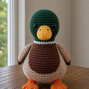 May include: A crochet mallard duck plush toy featuring a dark green head, a yellow beak, a white neck ring, a brown chest, light tan wings and body, and bright orange webbed feet. The duck has two small black plastic eyes and a round shape.
