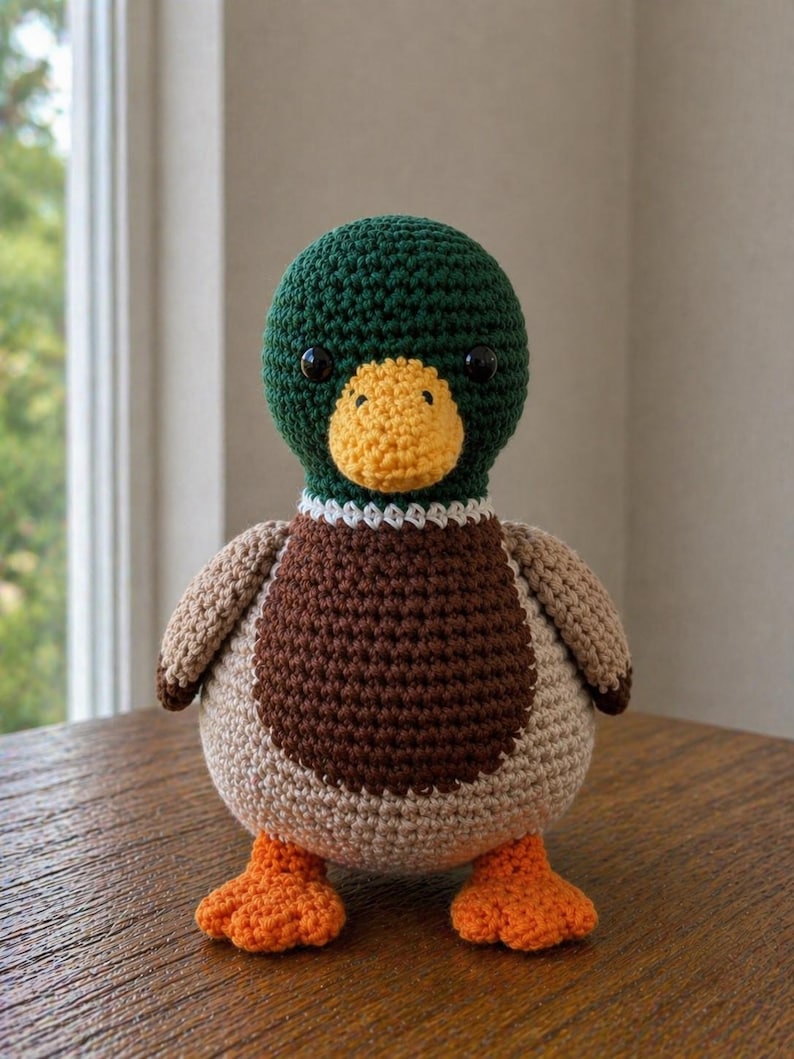 May include: A crochet mallard duck plush toy featuring a dark green head, a yellow beak, a white neck ring, a brown chest, light tan wings and body, and bright orange webbed feet. The duck has two small black plastic eyes and a round shape.