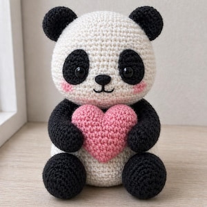 May include: A crocheted panda bear in black and white, clutching a pink heart. The panda has black eyes, ears, and limbs, with rosy cheeks. A charming, handmade soft toy, perfect as a gift or decorative item.