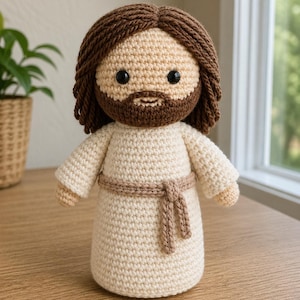 May include: A crocheted doll depicting a figure with brown hair and beard, wearing a cream-colored robe with a brown belt. The doll has black eyes and is standing upright. The background includes a plant in a woven pot and a window.
