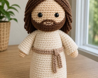 May include: A crocheted doll depicting a figure with brown hair and beard, wearing a cream-colored robe with a brown belt. The doll has black eyes and is standing upright. The background includes a plant in a woven pot and a window.