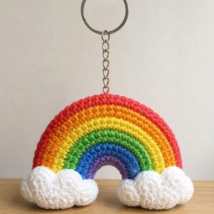 May include: A crocheted rainbow keychain with a silver-colored keyring and chain. The rainbow features bands of red, orange, yellow, green, blue, and purple. White, fluffy clouds are attached at the base of the rainbow.