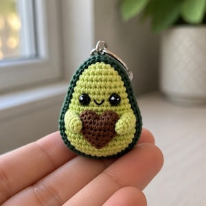 Könnte beinhalten: Ein handgehäkelter Avocado-Schlüsselanhänger. Die Avocado ist grün und gelb, mit einem dunkelgrünen Rand, einem lächelnden Gesicht und einem braunen Herz. Er hat einen silbernen Schlüsselring. Ein charmantes Accessoire.