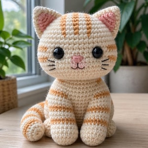 May include: A cream and orange crocheted cat figurine. The cat has large black eyes, a pink nose, and whiskers. The ears are lined with pink. The cat is sitting with its tail curled around its body. It is on a wooden surface.