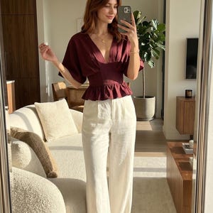 Peut inclure: Haut bordeaux à col en V avec manches courtes et taille péplum, associé à un pantalon blanc à jambes larges. La tenue se reflète dans un grand miroir, mettant en valeur une esthétique moderne et minimaliste. La pièce a une palette de couleurs neutres.