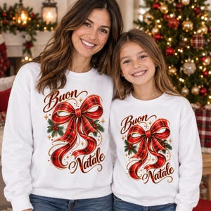 Può includere: Due felpe bianche a girocollo con una grande stampa di un fiocco a nastro scozzese rosso e nero. Il disegno include foglie di agrifoglio verdi con bacche rosse e la scritta "Buon Natale" in un carattere corsivo marrone con stelle dorate.