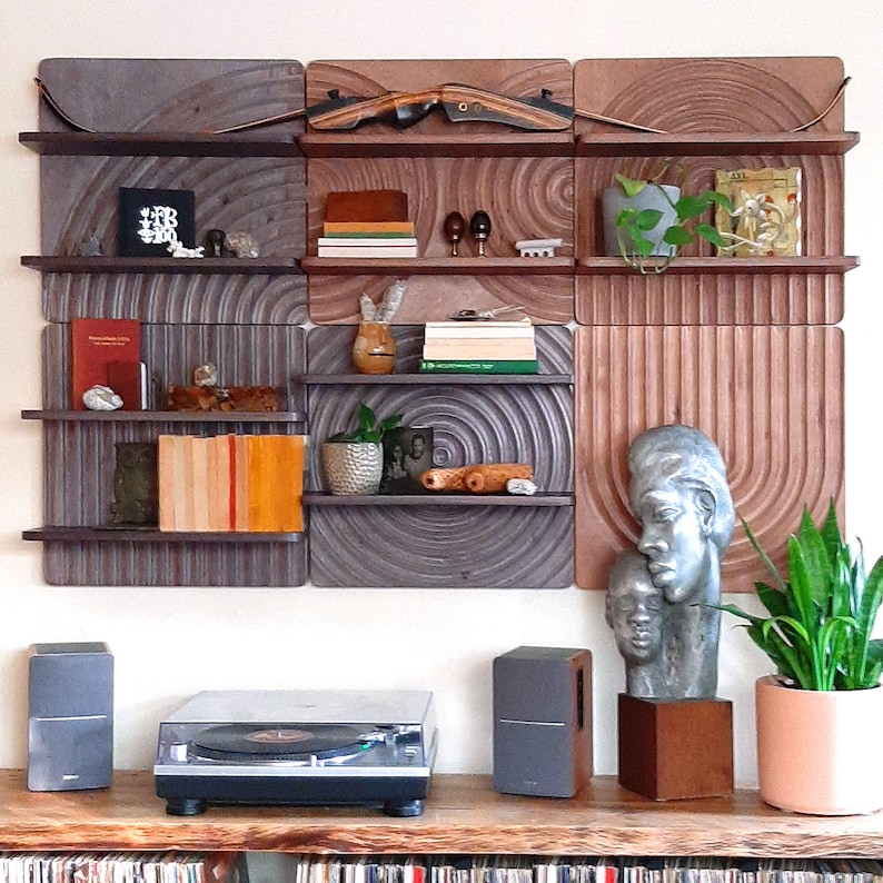 May include: A set of six wooden wall shelves with a circular pattern. The shelves are arranged in a grid pattern and are decorated with books, plants, and other decorative items. The shelves are mounted on a white wall. A record player and speakers are on a wooden table below the shelves.