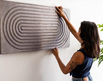 May include: A person hangs a large, gray, rectangular wall sculpture with a concentric circle design. The sculpture is made of wood and has a textured surface.