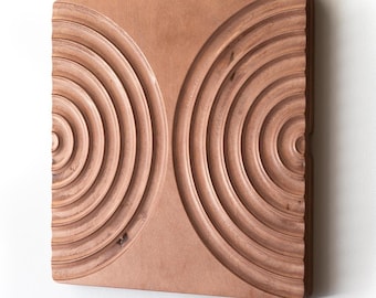 May include: A square wooden wall art piece with a carved design of two overlapping circles with concentric rings. The wood is a light brown color.