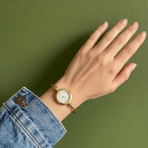Puede incluir: Un reloj de pulsera dorado con esfera blanca y números negros se lleva en la muñeca. El reloj tiene una correa fina y delicada. El fondo es verde liso y apagado. Una manga de chaqueta vaquera es visible en la esquina inferior izquierda.