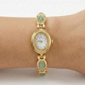 May include: Gold-toned wristwatch with an oval face and a white dial. The watch features a braided bezel and is adorned with oval-shaped green gemstones. The band is also gold-toned and has green gemstones.
