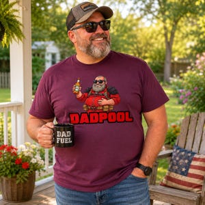 Könnte beinhalten: Ein kastanienbraunes Kurzarm-T-Shirt mit einer Grafik eines bärtigen Charakters in einem roten Anzug und Sonnenbrille, der eine Bierflasche hält, mit dem darunter gedruckten Text DADPOOL, getragen mit einer Baseballkappe und blauen Jeans.