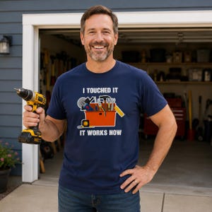 May include: Navy blue t-shirt with the text "I TOUCHED IT IT WORKS NOW" and a cartoon toolbox graphic. The toolbox graphic includes tools such as a saw, hammer, and screwdriver. The person is holding a yellow and black drill.